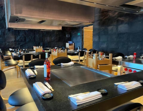 Modern teppanyaki restaurant interior with dark blue walls, wooden tables with built-in grills, place settings, and a yellow door in the background