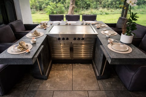 Configuración de comedor exterior moderno con parrilla central empotrada, encimeras de piedra, cubiertos y asientos negros con vista al césped verde