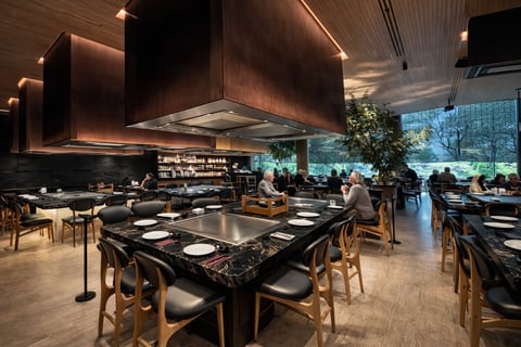 Interior de restaurante upscale moderno con mesas de comedor de mármol negro, sillas de madera, campana de ventilación de cobre, iluminación ambiental y ventanas del suelo al techo con vista a vegetación