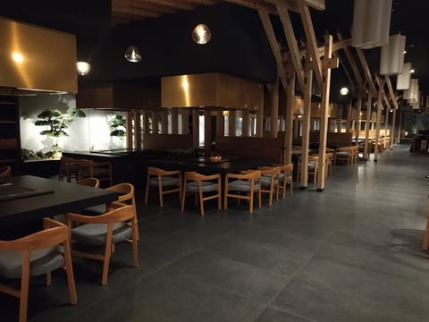 Interior de restaurante moderno con sillas de madera, pared divisoria curva de bronce, lámparas colgantes y suelos de hormigón gris por la noche
