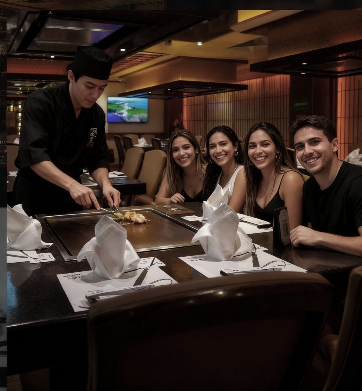 A chef serves food to four smiling guests seated at a teppanyaki grill table in an upscale restaurant