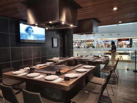 Modern sushi bar restaurant with central wooden counter, place settings, flat-screen TV, and open kitchen visible in background