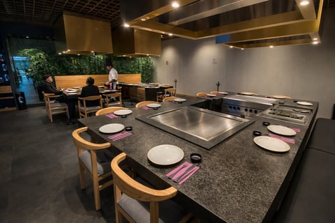 Restaurante teppanyaki moderno con dos grandes encimeras de plancha, sillas curvadas de madera, configuraciones blancas y decoración de pared verde con plantas bajo luces cálidas del techo