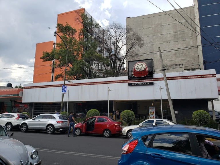 Street view of a restaurant storefront with parked cars, orange and gray buildings, and vegetation