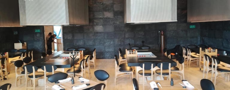 Modern restaurant dining room with wooden chairs, set tables, dark blue tiled backsplash, and white pendant lights