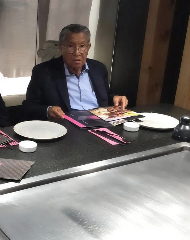 Older man in dark blazer and light blue shirt seated at a dining table with place settings and pink napkins