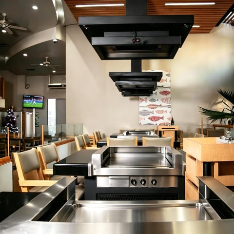 Modern restaurant kitchen with stainless steel cooking equipment, black ventilation hood, wood furnishings, and contemporary ceiling design