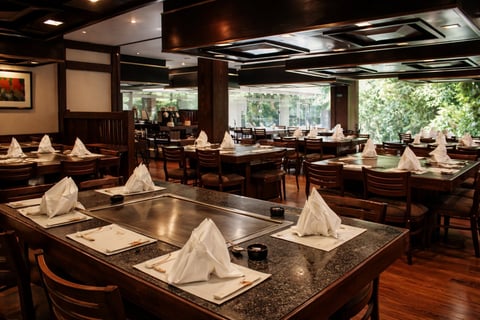 Salón de comedor de restaurante upscale con mobiliario de madera oscura, mesas con tapa de granito y configuraciones con servilletas blancas, grandes ventanas con vista a vegetación