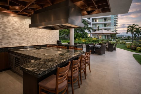 Cocina exterior moderna con isla de granito, asientos de bar, electrodomésticos de acero inoxidable y vista a edificios residenciales y palmeras
